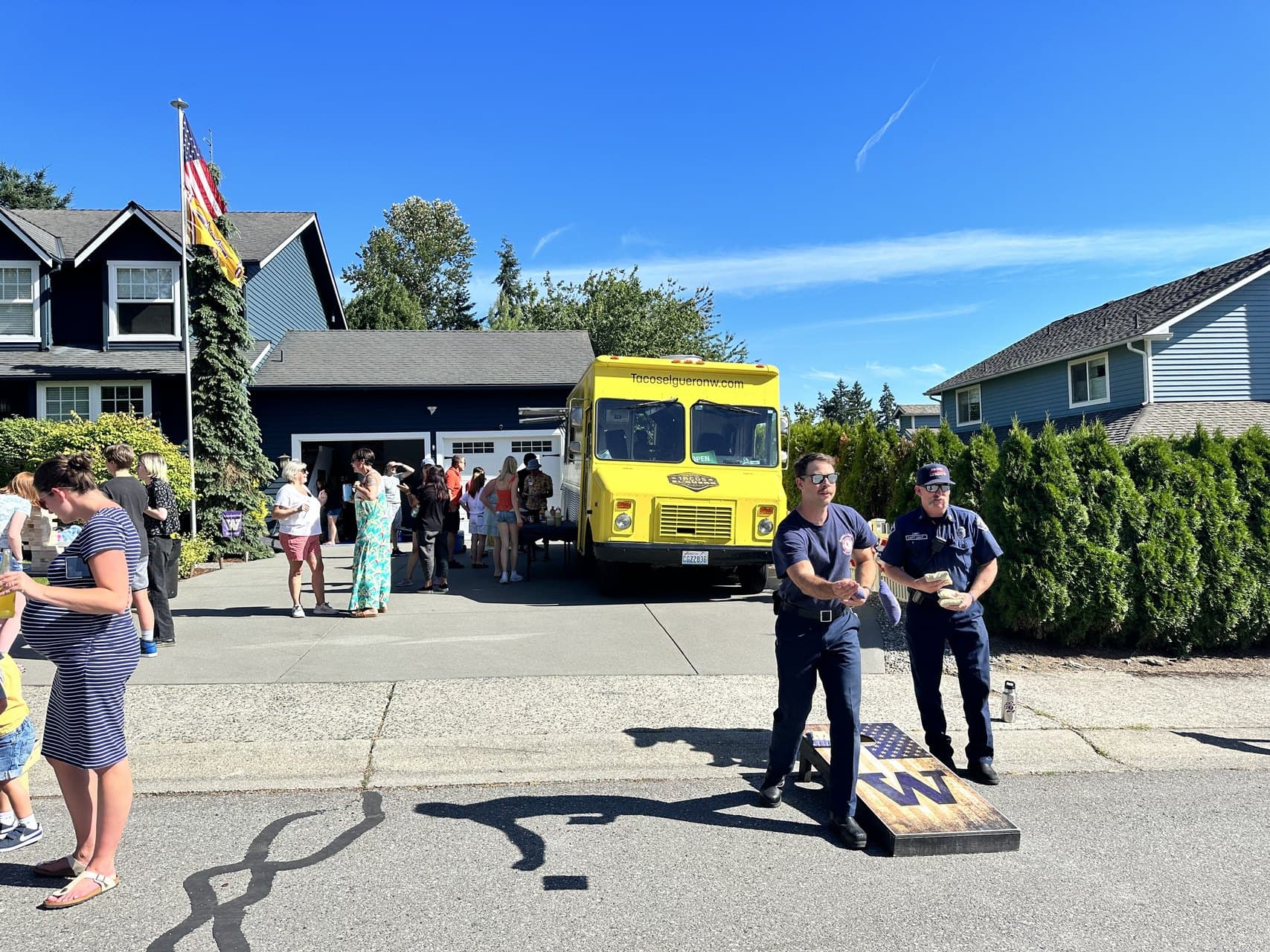 Two Eastside Fire firefighters playing cornhole in the street at a Duvall block party, with a yellow taco truck in the driveway behind them